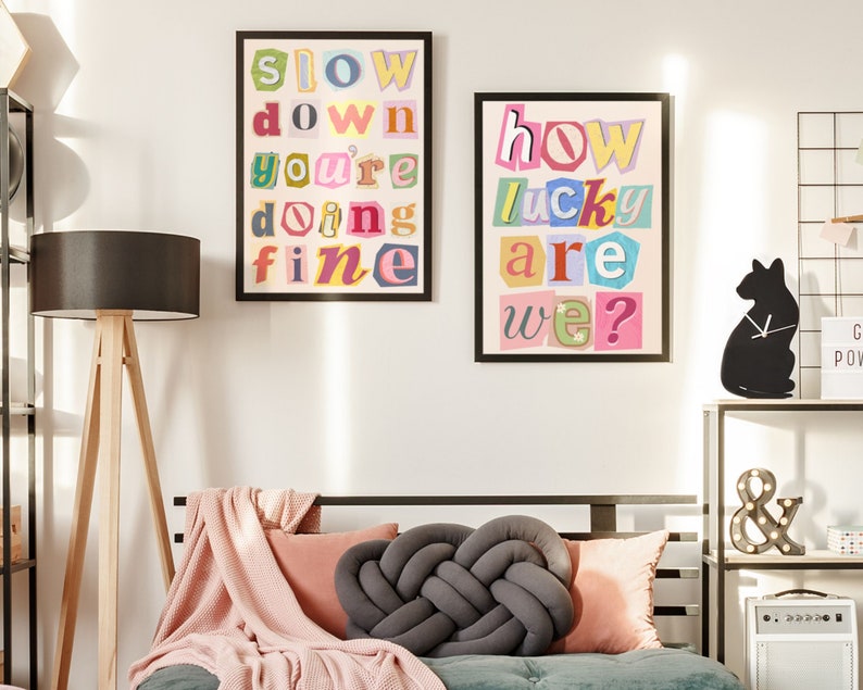 Op de afbeelding: Twee ingelijste prints met kleurrijke uitgesneden letters die "slow down you're doing fine" en "how lucky are we?" spellen, hangen aan een witte muur boven een grijze bank met een roze deken en een grijs geknoopt kussen.