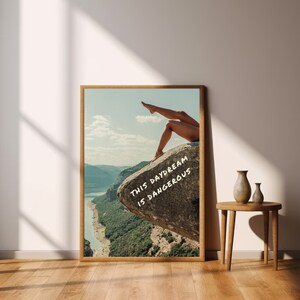 Peut inclure: Une affiche encadr&eacute;e repr&eacute;sentant un paysage avec les jambes d'une personne d&eacute;passant d'un rocher. Le texte "THIS DAYDREAM IS DANGEROUS" est &eacute;crit sur le rocher. L'affiche est dans un cadre en bois, avec une rivi&egrave;re et des montagnes.