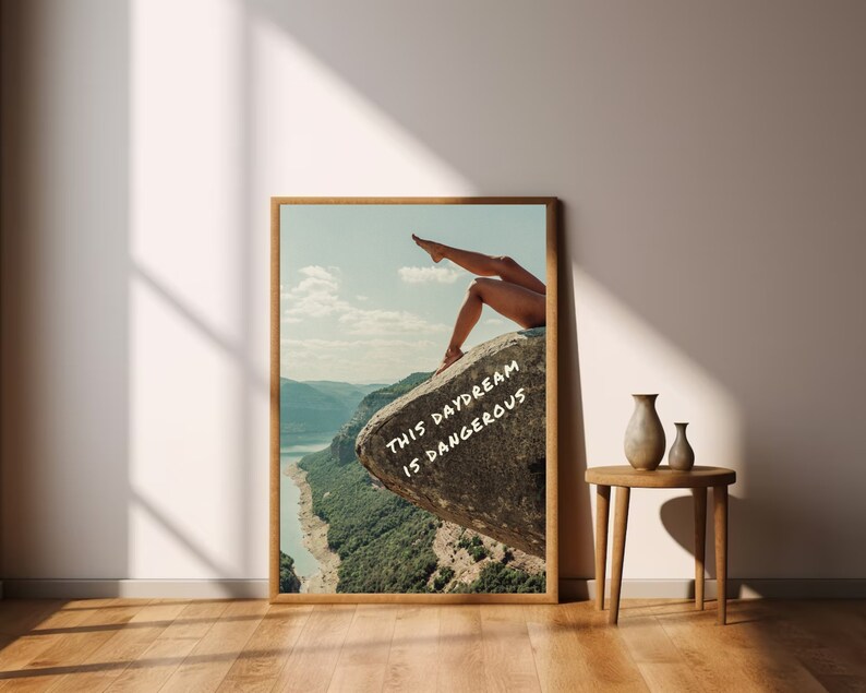Peut inclure: Une affiche encadr&eacute;e repr&eacute;sentant un paysage avec les jambes d'une personne d&eacute;passant d'un rocher. Le texte "THIS DAYDREAM IS DANGEROUS" est &eacute;crit sur le rocher. L'affiche est dans un cadre en bois, avec une rivi&egrave;re et des montagnes.
