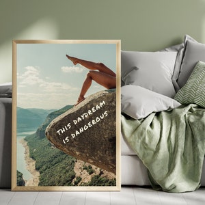 Peut inclure: Impression d'art encadr&eacute;e repr&eacute;sentant un paysage avec les jambes d'une personne sur un rocher. L'&oelig;uvre d'art comprend le texte "THIS DAYDREAM IS DANGEROUS" en blanc. L'impression est expos&eacute;e dans une pi&egrave;ce avec un mur vert et un canap&eacute; gris.