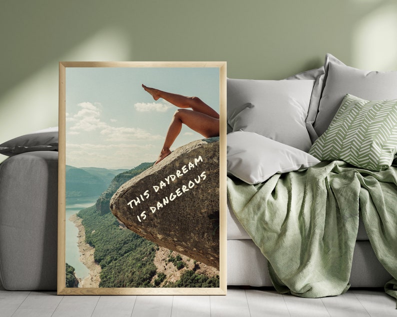 Peut inclure: Impression d'art encadr&eacute;e repr&eacute;sentant un paysage avec les jambes d'une personne sur un rocher. L'&oelig;uvre d'art comprend le texte "THIS DAYDREAM IS DANGEROUS" en blanc. L'impression est expos&eacute;e dans une pi&egrave;ce avec un mur vert et un canap&eacute; gris.