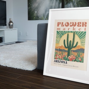 Op de afbeelding: Een wit ingelijste poster met een vintage ontwerp met een Saguaro-cactus in een woestijnlandschap. De poster luidt "Flower Market Arizona State Flower - Saguaro Cactus Blossom".