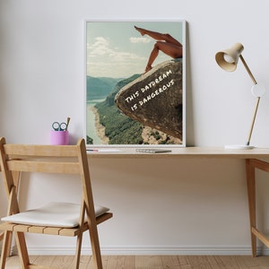 Peut inclure: Impression d'art encadr&eacute;e avec des jambes sur un rocher surplombant un paysage. Le texte "THIS DAYDREAM IS DANGEROUS" est &eacute;crit en blanc. L'&oelig;uvre est sur un bureau en bois avec une lampe et un pot &agrave; crayons rose.