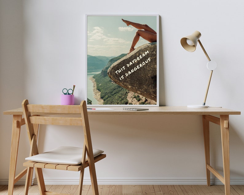 Peut inclure: Impression d'art encadr&eacute;e avec des jambes sur un rocher surplombant un paysage. Le texte "THIS DAYDREAM IS DANGEROUS" est &eacute;crit en blanc. L'&oelig;uvre est sur un bureau en bois avec une lampe et un pot &agrave; crayons rose.