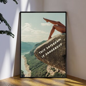 Peut inclure: Impression d'art encadr&eacute;e repr&eacute;sentant un paysage avec une &eacute;tendue d'eau et des montagnes. L'image comprend les jambes d'une personne d&eacute;passant d'un rocher avec le texte "THIS DAYDREAM IS DANGEROUS" en blanc.