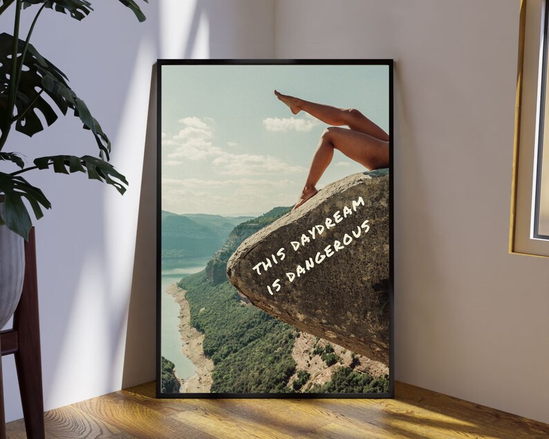Peut inclure: Impression d'art encadr&eacute;e repr&eacute;sentant un paysage avec une &eacute;tendue d'eau et des montagnes. L'image comprend les jambes d'une personne d&eacute;passant d'un rocher avec le texte "THIS DAYDREAM IS DANGEROUS" en blanc.