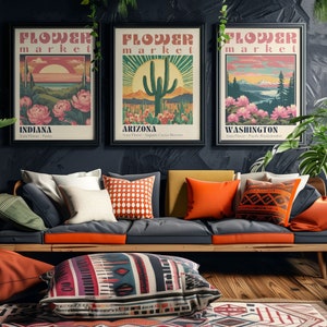 Może przedstawiać: Trzy plakaty w stylu vintage z tekstem "Flower Market" i nazwami r&oacute;żnych stan&oacute;w: Indiana, Arizona i Waszyngton. Plakaty przedstawiają ilustracje kwiat&oacute;w, kaktusa i g&oacute;rskiego krajobrazu. Plakaty wiszą na ciemnej ścianie nad sofą z pomarańczowymi i wzorzystymi poduszkami.