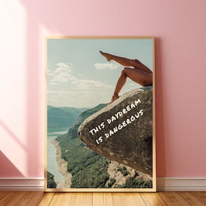 Peut inclure: Une affiche encadr&eacute;e repr&eacute;sentant un paysage avec les jambes d'une personne sur un rocher. L'affiche contient le texte "THIS DAYDREAM IS DANGEROUS" en blanc. L'&oelig;uvre est pr&eacute;sent&eacute;e sur un mur rose.