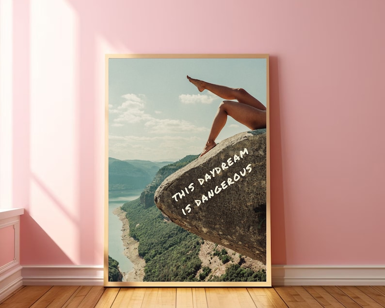 Peut inclure: Une affiche encadr&eacute;e repr&eacute;sentant un paysage avec les jambes d'une personne sur un rocher. L'affiche contient le texte "THIS DAYDREAM IS DANGEROUS" en blanc. L'&oelig;uvre est pr&eacute;sent&eacute;e sur un mur rose.