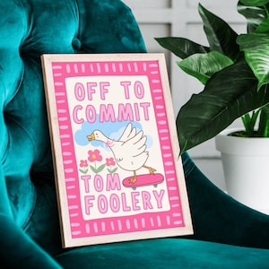May include: A framed art print with a pink border and the words "OFF TO COMMIT TOM FOOLERY." The artwork features a cartoon goose riding a skateboard, with flowers and clouds. The frame is a light wood colour.