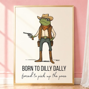 May include: A framed wall art print featuring a green frog dressed as a cowboy in a tan hat, brown vest, and neckerchief, holding a revolver. Below the illustration, the text reads: BORN TO DILLY DALLY forced to pick up the pace.