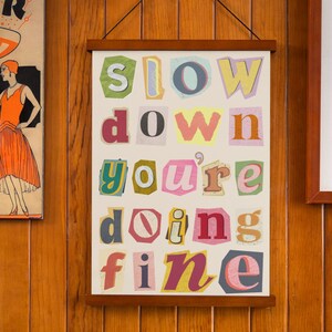 Puede incluir: Una impresión enmarcada con las palabras "slow down you're doing fine" en letras de colores recortadas. Las letras están dispuestas en un patrón aleatorio sobre un fondo blanco.