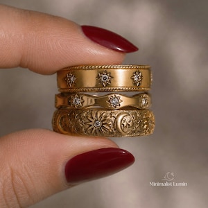 May include: A stack of three gold-toned rings. The top ring features a rope-textured edge and star motifs with small clear stones. The middle ring has a wavy edge and star engravings. The bottom ring displays a textured celestial sun and moon design.