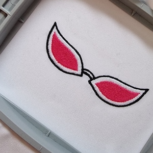 Può includere: Disegno ricamato di due occhi rosa con contorni neri su tessuto bianco in un telaio da ricamo.