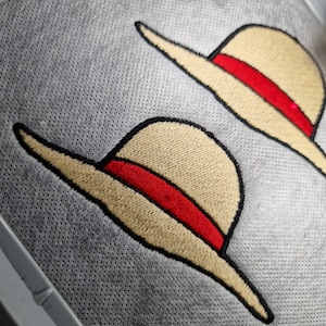 Può includere: Due cappelli beige ricamati con bande rosse su una superficie grigia testurizzata.