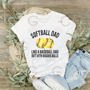 Może przedstawiać: Biała koszulka z czarnym tekstem, kt&oacute;ry brzmi "Softball Dad Like a Baseball Dad But With Bigger Balls". Dwie ż&oacute;łte grafiki baseballa znajdują się nad tekstem.