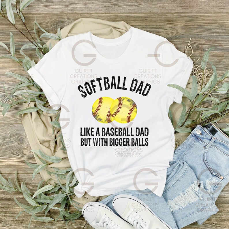 Może przedstawiać: Biała koszulka z czarnym tekstem, kt&oacute;ry brzmi "Softball Dad Like a Baseball Dad But With Bigger Balls". Dwie ż&oacute;łte grafiki baseballa znajdują się nad tekstem.