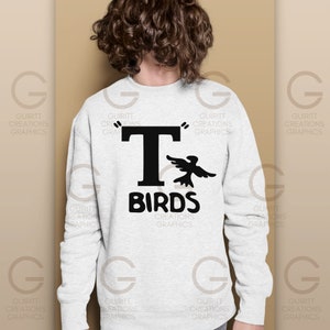 May include: A white sweatshirt with a black graphic of a bird flying next to a large letter "T" and the text "BIRDS" underneath. The text "GUIRITT CREATIONS GRAPHICS" is printed on the bottom of the sweatshirt.