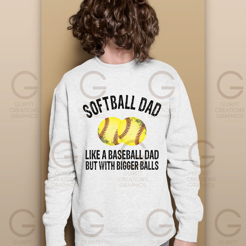 Może przedstawiać: Biała bluza z nadrukiem "Softball Dad Like a Baseball Dad But With Bigger Balls". Dwa ż&oacute;łte grafiki piłek softball znajdują się nad tekstem.