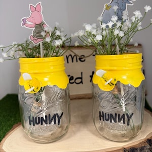 May include: Two clear glass mason jars with yellow painted rims resembling dripping honey. Each jar features the word HUNNY in black lettering, a small fuzzy bee embellishment, and white baby's breath flowers with character cutouts.