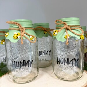 May include: Clear glass Ball Mason jars with green lids and the word "HUNNY" in black. Each jar is decorated with a rope bow and bee embellishments. The jars are arranged on a wooden slice.