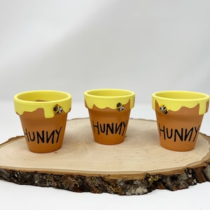 May include: Three small terracotta pots with yellow paint and a dripping honey design. Each pot has a black bee painted on the side and the word "Hunny" written in black.