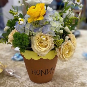May include: A yellow, white and blue flower arrangement in a terracotta pot with the word "Hunny" written on it. The pot has a yellow scalloped rim.