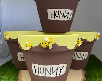 May include: Three brown terracotta flower pots with yellow and white paint accents. Each pot has a small yellow and black bee figurine attached to the rim. The pots are stacked on top of each other and the bottom pot is sitting on a wooden slice. The pots are labeled "Hunny".
