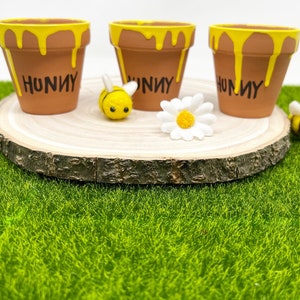 May include: Three brown terracotta flower pots with yellow paint dripping from the top, each with the word "HUNNY" in black. A felt bee and a white daisy sit on a wooden slice, all on a green surface.