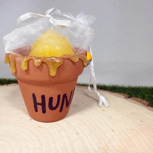 May include: A terracotta-coloured clay pot featuring the word HUNY painted in dark purple letters. The rim is decorated with a yellow, glittery, dripping honey-like substance. A yellow object sits inside a white, sheer, drawstring organza bag.