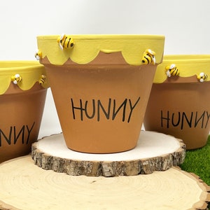 Peut inclure: Trois pots de fleurs en terre cuite avec des accents d'abeilles jaunes et blanches. Les pots sont décorés d'un motif de nid d'abeille jaune et du mot "Hunny" écrit en noir. Les pots sont posés sur une tranche de bois.