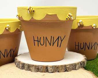 May include: Three terracotta flower pots with yellow and white bee accents. The pots are decorated with a yellow honeycomb design and the word "Hunny" written in black. The pots are sitting on a wooden slice.