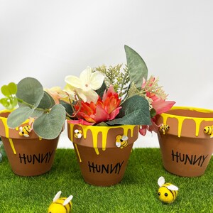 May include: Three terracotta flower pots with yellow dripping paint and the word "Hunny" written on them. Each pot has a small bee figurine on the rim. The pots are sitting on a green artificial grass surface.