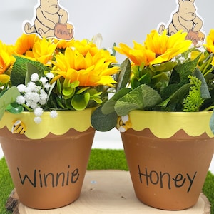 Peut inclure: Deux pots de fleurs en terre cuite avec des bords festonnés jaunes, remplis de tournesols et de verdure. Chaque pot porte une figurine de Winnie l'ourson et les mots "Winnie" et "Honey". Des abeilles décoratives sont attachées aux pots.