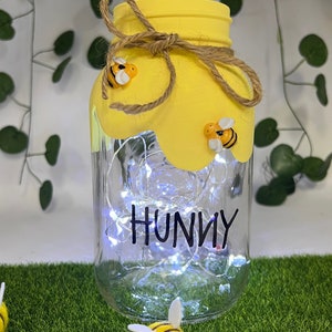 May include: A yellow mason jar with a string bow and bee decorations. The jar has white fairy lights inside and the word "Hunny" written on the front.