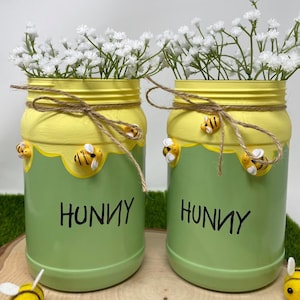 May include: Two green and yellow painted jars with the word "HUNNY" in black, decorated with bee accents and twine. Each jar holds white flowers. Decorative bee figurines are placed near the jars.