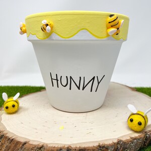 May include: White ceramic planter with a yellow rim and dripping honey design. The planter has the word "Hunny" written on the front and is decorated with small yellow and black bee figurines.