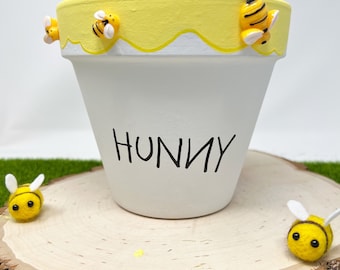 May include: White ceramic planter with a yellow rim and dripping honey design. The planter has the word "Hunny" written on the front and is decorated with small yellow and black bee figurines.