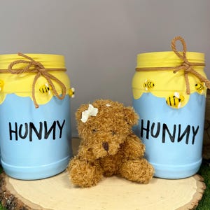 May include: Two painted mason jars with yellow lids and blue bases, adorned with bee designs and the word "HUNNY". A small teddy bear sits between the jars on a wooden slice, creating a whimsical, themed display.