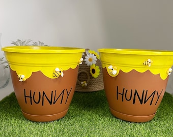 May include: Two brown and yellow plant pots with a honey-drip design at the rim. Each pot features the word HUNNY in black lettering and small, three-dimensional bee figures attached to the exterior. The pots have a matching saucer base.