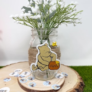 May include: A glass mason jar with a Winnie the Pooh sticker on it, filled with white baby's breath flowers. The jar is tied with twine and has a small bee sticker on it. The jar is sitting on a wooden slice with Winnie the Pooh confetti scattered around it.
