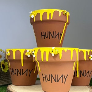 May include: Four brown terracotta flower pots with yellow paint dripping from the rims, resembling honey. Each pot has the word "HUNNY" in black and is decorated with small bee figurines. The pots are arranged on a wooden slice.