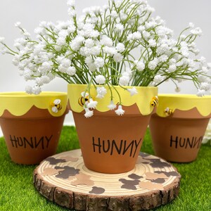 Peut inclure: Trois pots de fleurs en terre cuite peints en jaune et brun, décorés d'abeilles et du mot "Hunny". Les pots sont remplis de fleurs de gypsophile blanches et sont posés sur une tranche de bois.