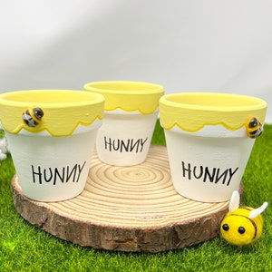 Peut inclure: Trois petits pots en céramique blanche avec des bords jaunes et un motif de miel. Chaque pot porte le mot "Hunny" écrit sur le devant. Les pots sont posés sur une tranche de bois et entourés d'herbe verte. Une petite abeille en feutre jaune avec des rayures noires est assise sur la tranche de bois.