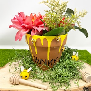 May include: A terracotta flower pot with a yellow honey drip design and the word "Hunny" written on it. The pot is decorated with small yellow bees and contains pink flowers and green foliage.