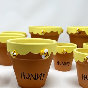 May include: Set of seven terracotta flower pots painted with a yellow honey drip design and a bee accent. The pots are labeled "Hunny".