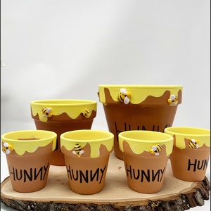 May include: A collection of brown terracotta flower pots with a yellow honey-like paint design around the rims. Each pot has the word "HUNNY" in black and small bee embellishments. The pots are arranged on a wooden slice.