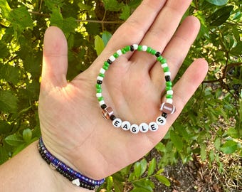 May include: A beaded bracelet with the word "EAGLES" spelled out in white letter beads. The bracelet features green, black, and white beads, with two football-shaped beads. Additional bracelets are visible in the background.