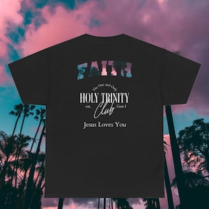 May include: Black t-shirt featuring the word "FAITH" in a colourful, tropical-themed font. The shirt also displays "HOLY TRINITY CLUB" and "Jesus Loves You" in white text. The background shows a sunset and palm trees, creating a vibrant scene.