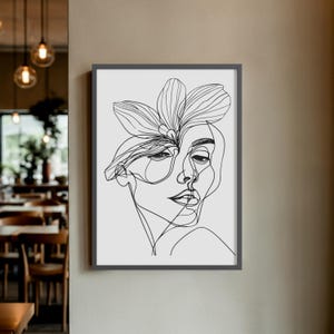 May include: A framed black and white line art print. The artwork shows a woman's face with a flower in her hair. The minimalist design uses continuous lines, creating a modern and elegant aesthetic. The print is displayed on a light grey background.